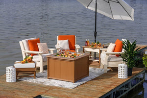 Three white poly patio chairs with orange pillows around a cedar fire pit table with food on a lakeside dock
