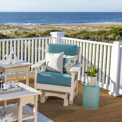 White poly glider chair with teal cushions and floral pillows on an oceanfront deck beside white side tables