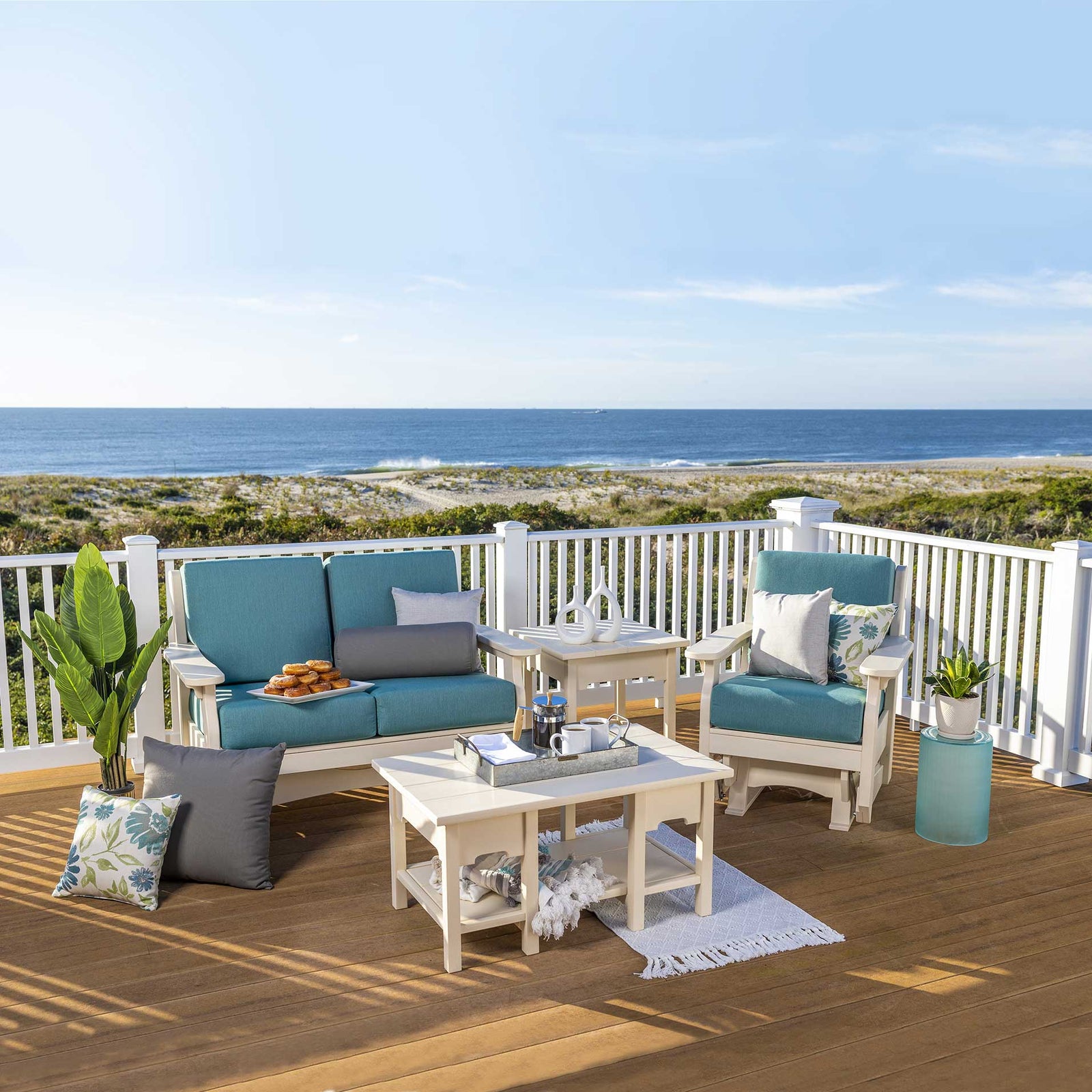 White poly glider loveseat, glider chair, side table and coffee table with teal cushions on a beachfront deck