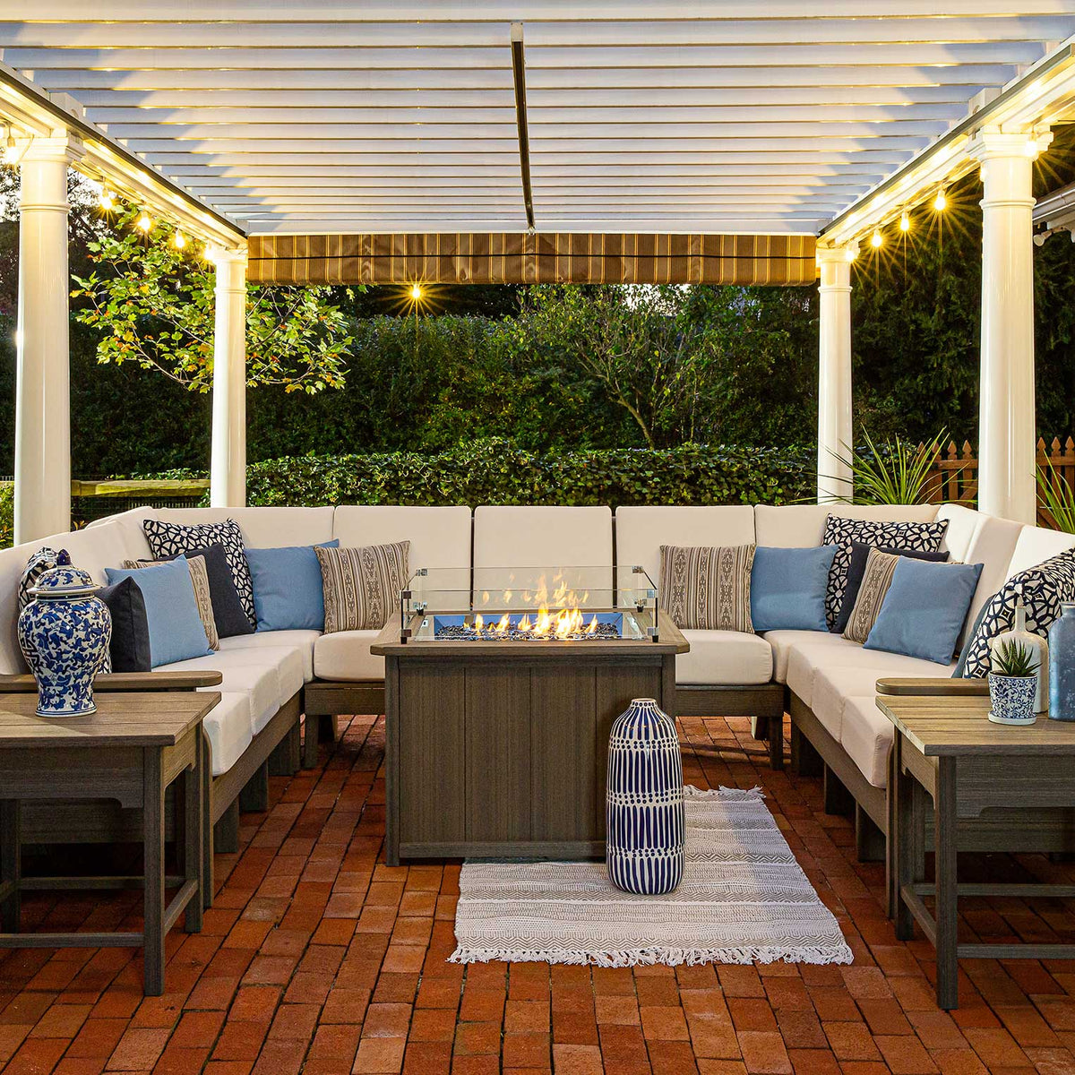 Large U shaped poly sectional with cream cushions and blue pillows around a lit fire table under a lit pergola
