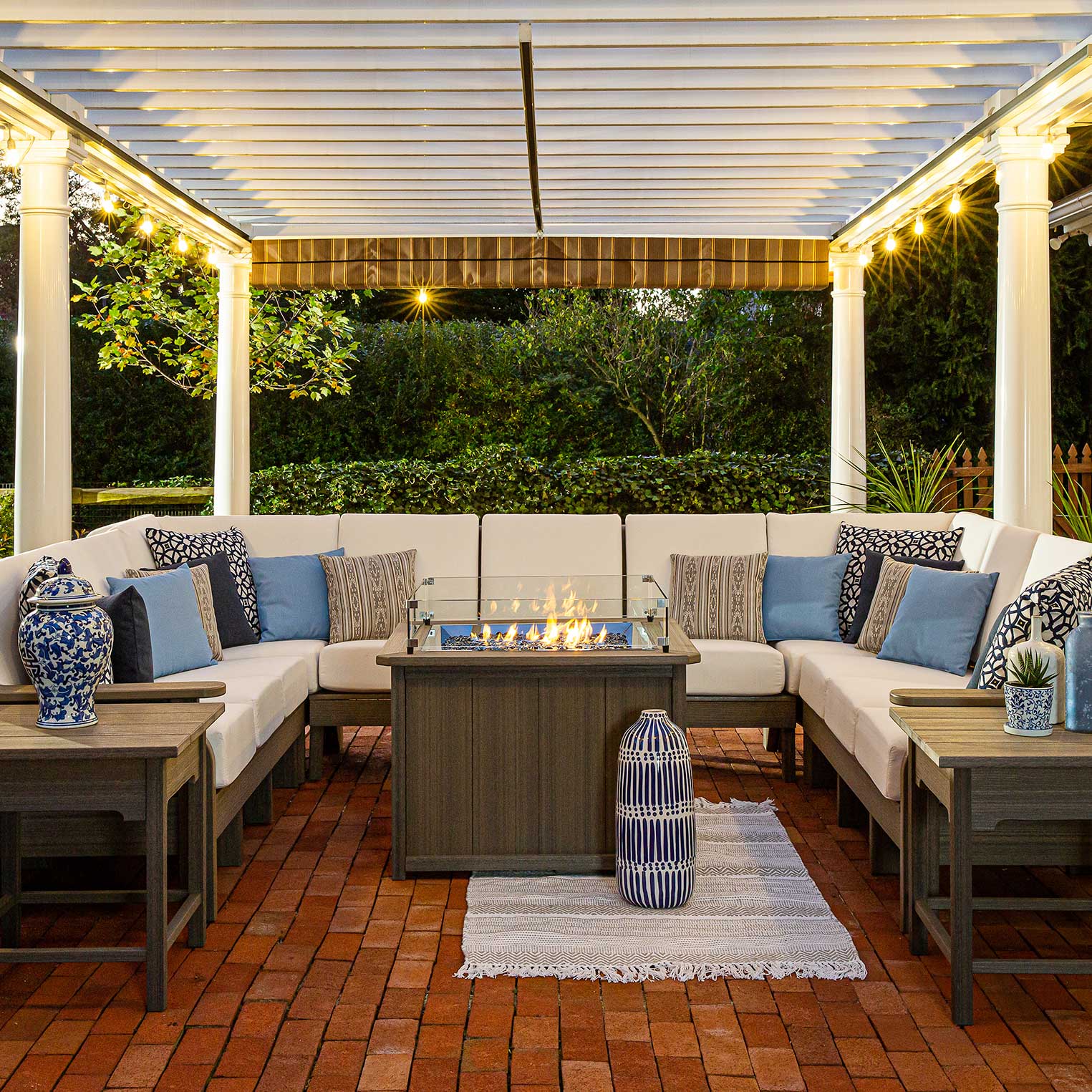 Large U shaped poly sectional with cream cushions and blue pillows around a lit fire table under a lit pergola