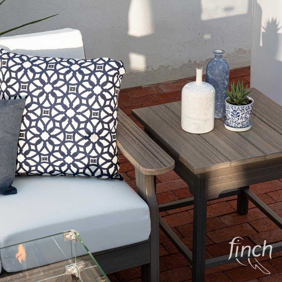 Weathered gray poly side table with vases and a potted plant beside a white cushioned chair and a lit fire table