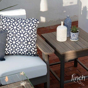 Weathered gray poly side table with vases and a potted plant beside a white cushioned chair and a lit fire table