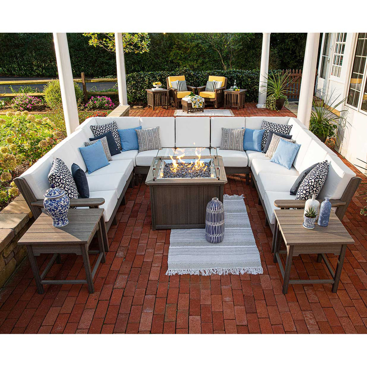 Evening patio with a U shaped sectional, white cushions, patterned pillows and a burning fire table on brick pavers