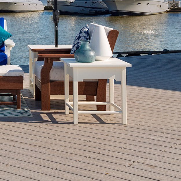 White poly end table on a marina dock next to a mahogany patio chair with white cushions and moored boats behind