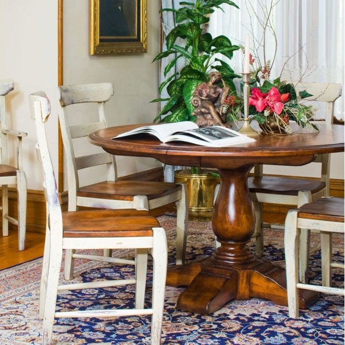 Round cherry pedestal dining table with a turned urn base and distressed cream ladder back chairs on a blue rug
