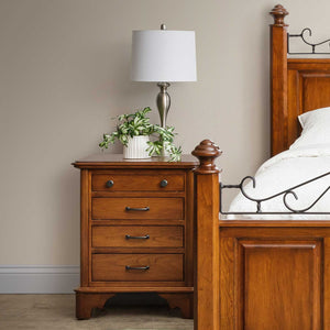 Cherry four drawer night stand with bronze pulls and a scalloped bracket base beside an iron scroll bed