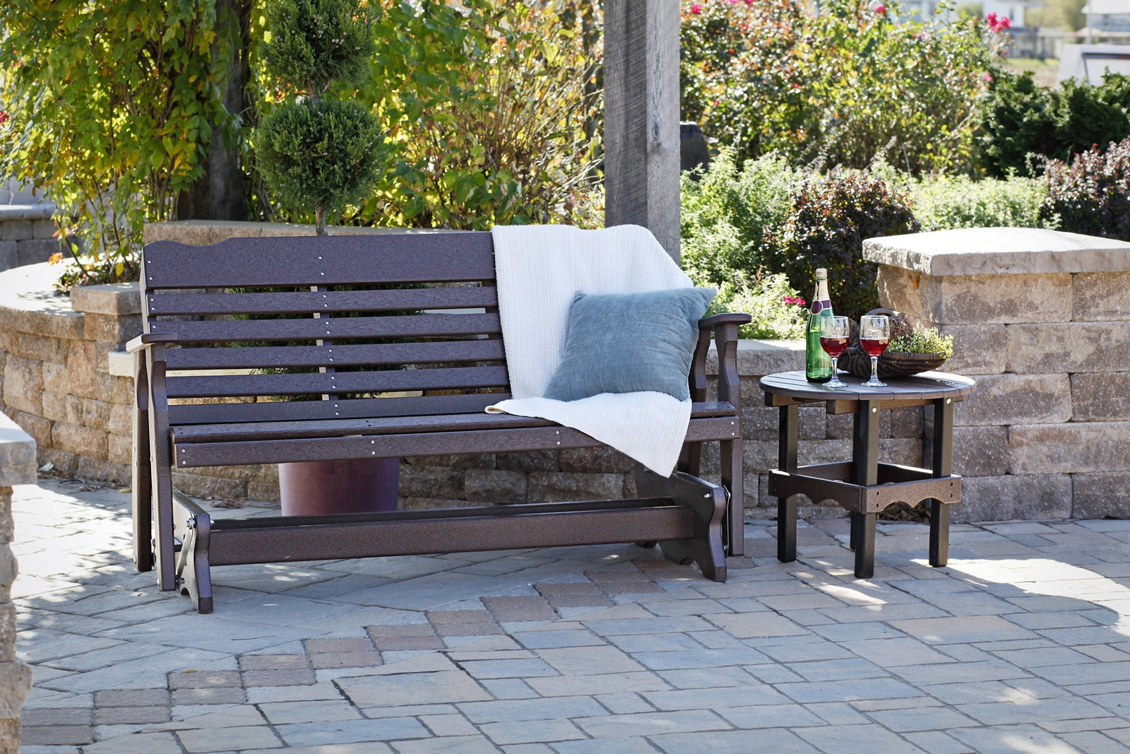 Dark brown poly West Chester glider bench with a throw and pillow, paired with a round side table on pavers