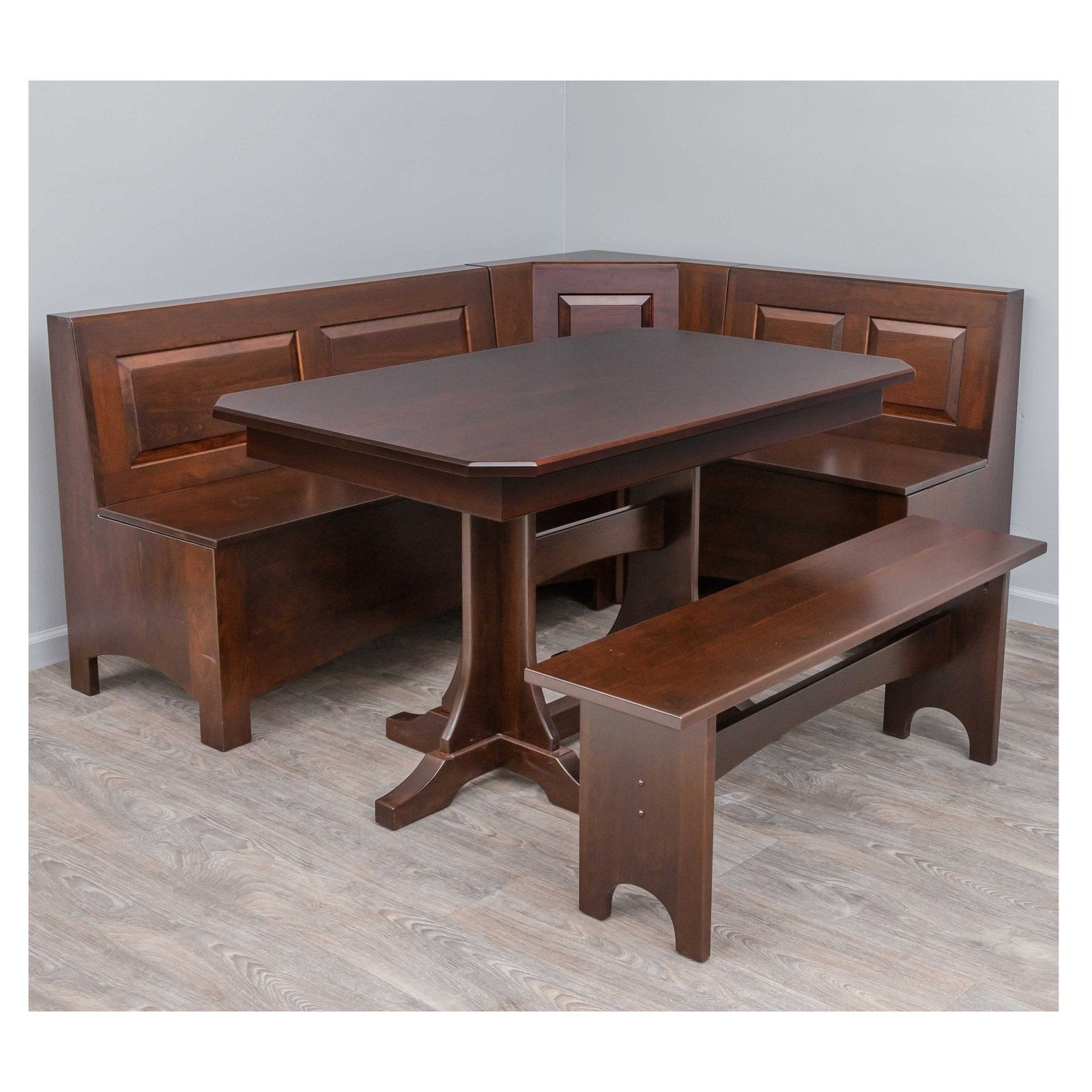 Gray stained corner breakfast nook set with raised panel bench backs, storage seating, pedestal table and matching bench in a showroom