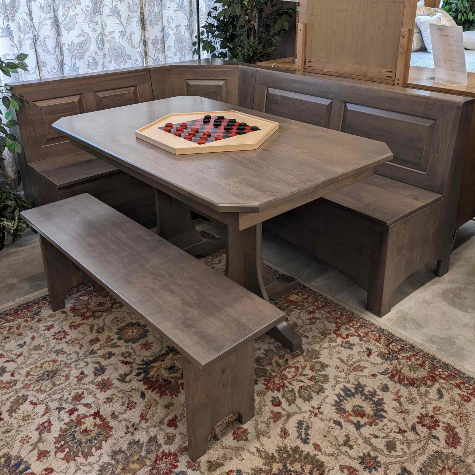 Gray stained corner breakfast nook set with raised panel bench backs, storage seating, pedestal table and matching bench in a showroom