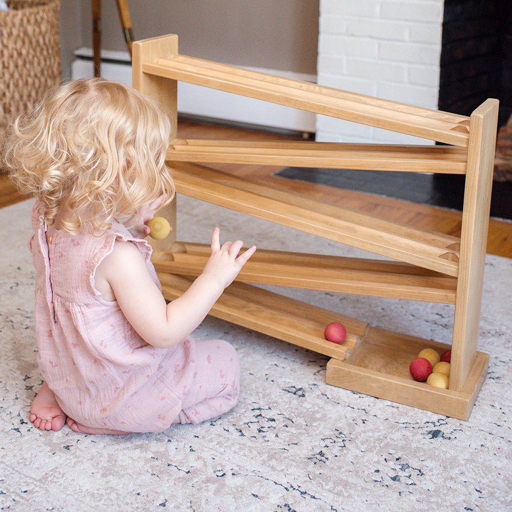 Toddler in a pink outfit kneeling to roll a wooden ball down a wooden ball roller racetrack on a rug