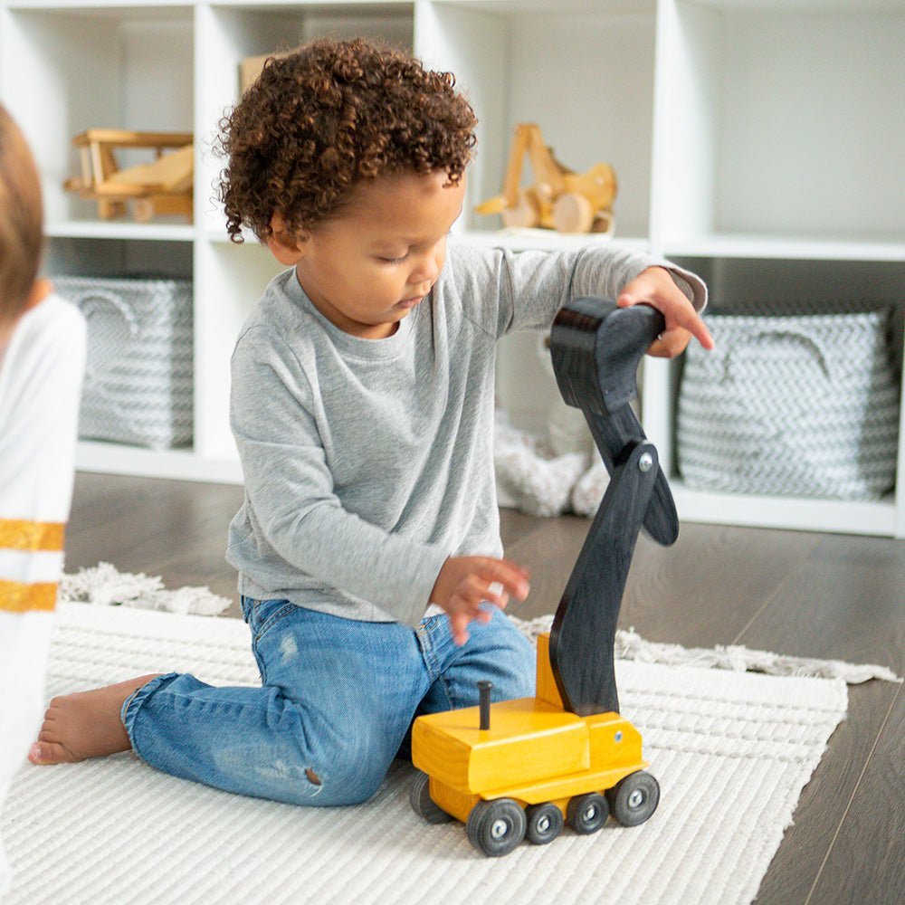 Toddler kneeling on a play rug swinging the black boom of a yellow painted wooden toy excavator in a bright playroom