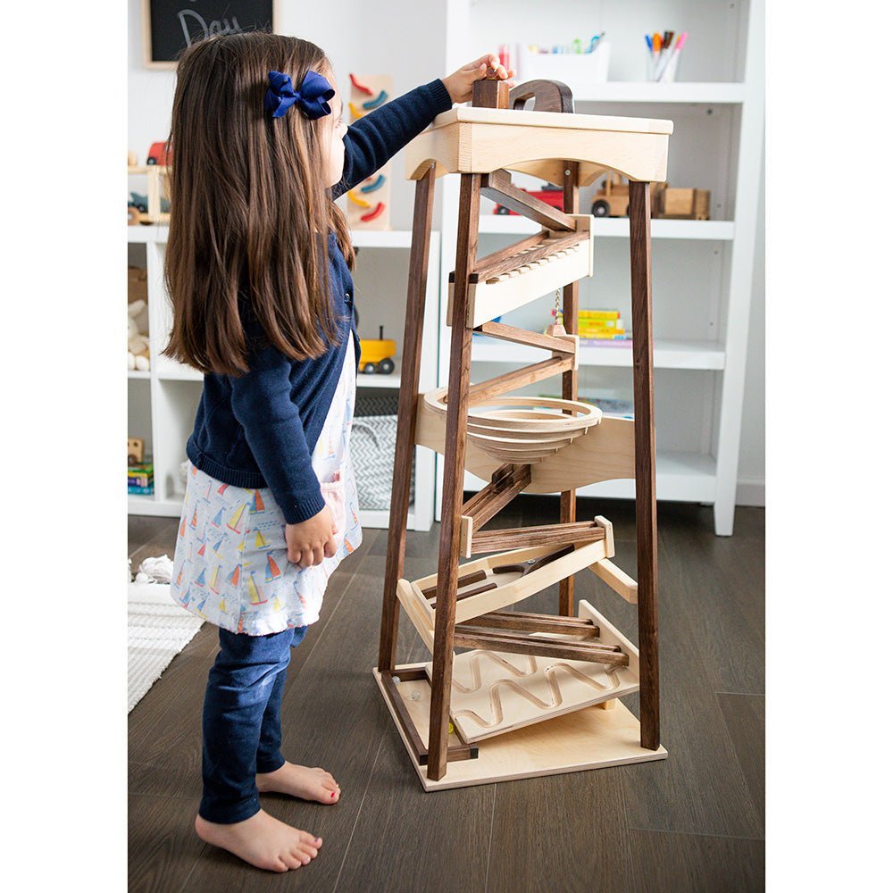 Girl dropping a marble into the top of a tall two-tone wooden marble pyramid tower in a bright playroom