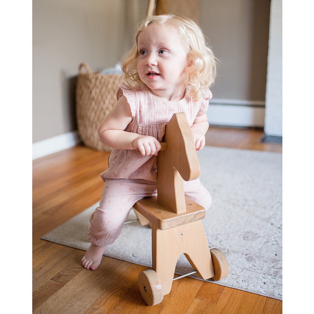 Toddler girl in pink straddling a natural wood ride-on horse with handle pegs and wheels on a living room rug