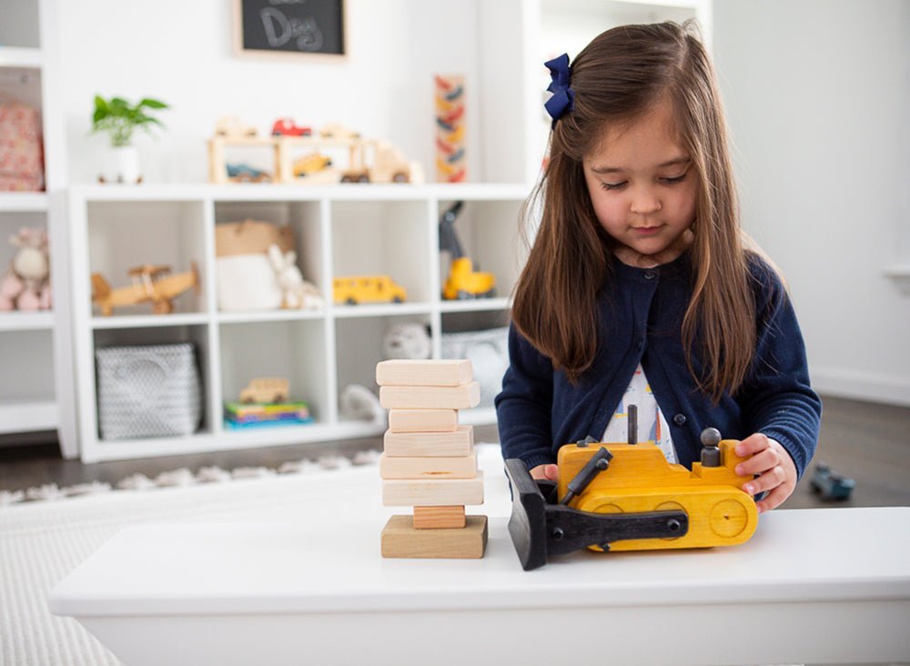 Girl pushing a yellow wooden toy bulldozer with black blade toward a stack of wooden building blocks