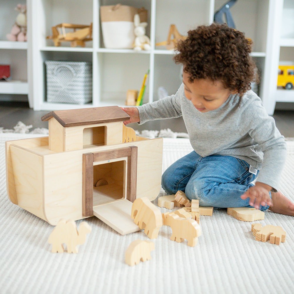 Toddler kneeling on a rug lifting the roof off a wooden Noah's ark toy with animal figures scattered around