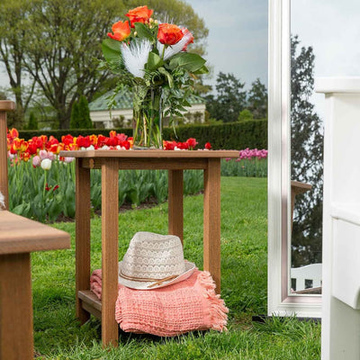 Teak poly outdoor side table holding a vase of roses, with a hat and blanket on the lower shelf