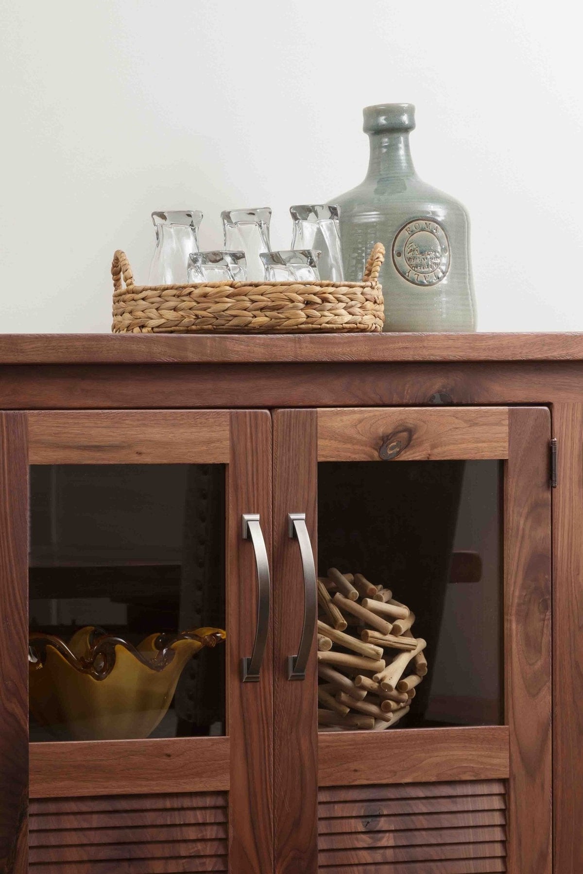 Detail of walnut sideboard glass doors with brushed nickel bar pulls, louvered lower panels and decor styled on the plank top
