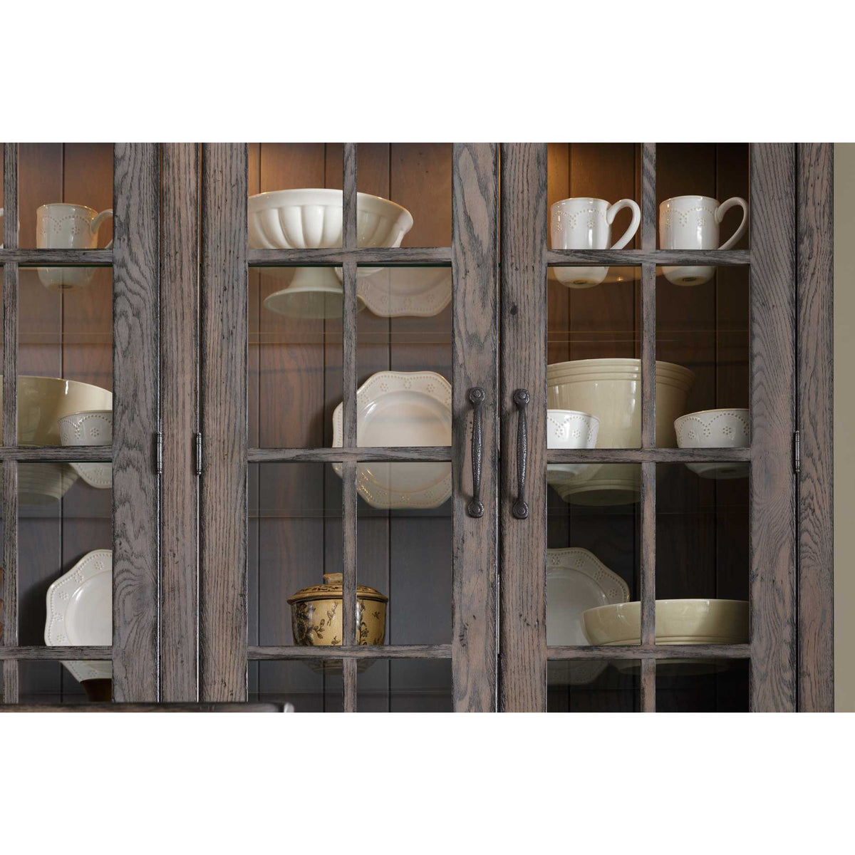 Lit china cabinet interior seen through mullioned glass doors, filled with cream dishes, mugs and bowls on glass shelves