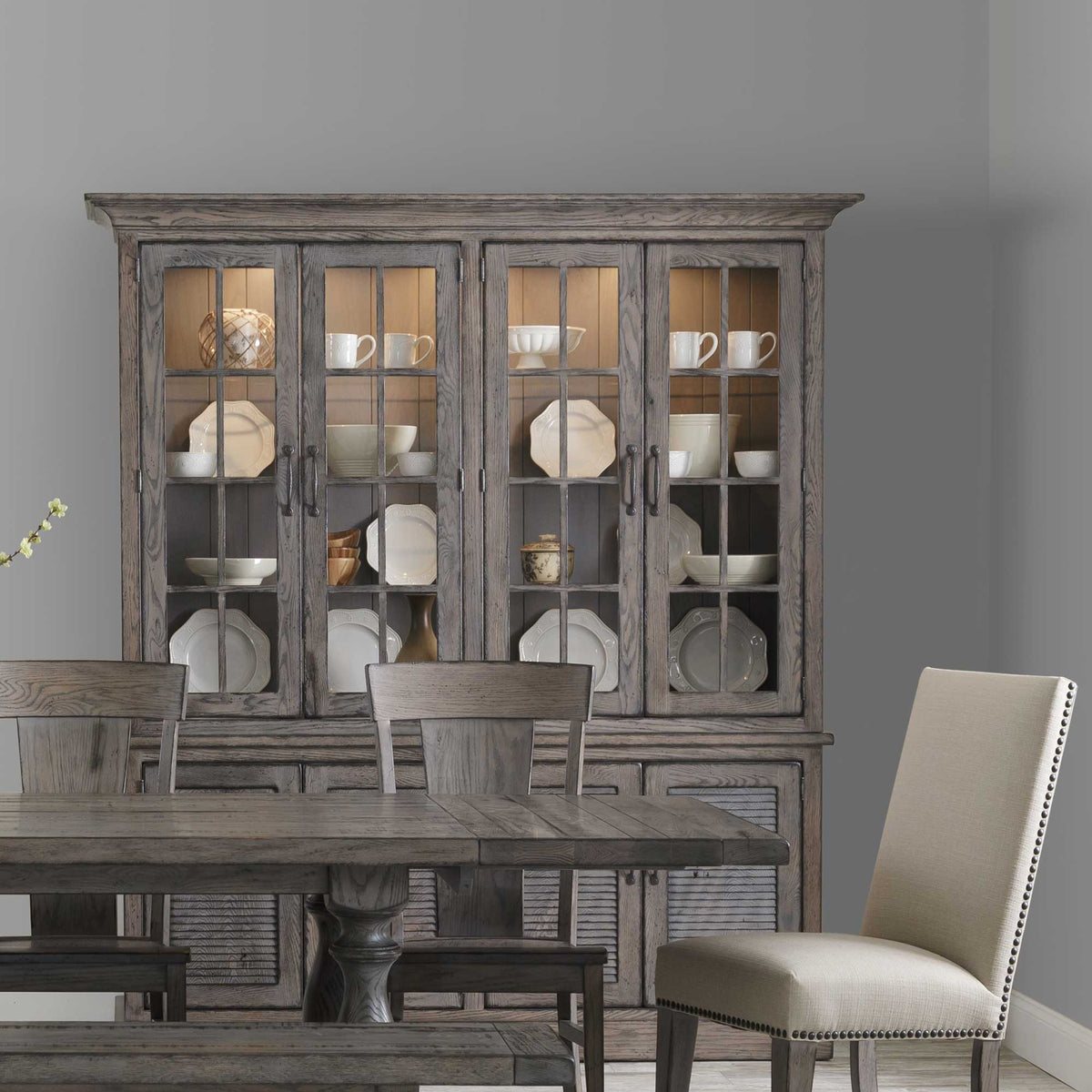 Driftwood gray four-door china cabinet filled with white dishware above a dining table with upholstered chairs