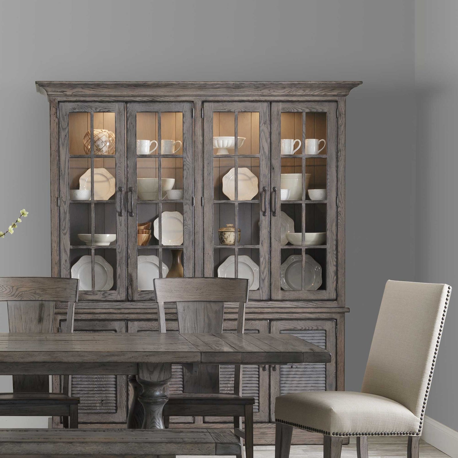 Driftwood gray four-door china cabinet filled with white dishware above a dining table with upholstered chairs