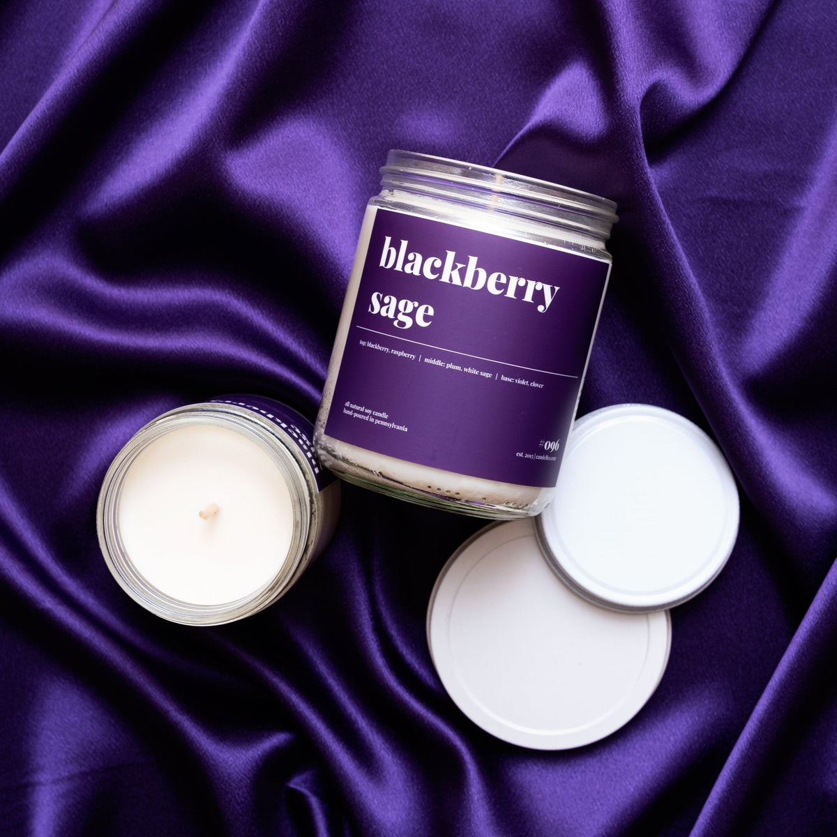 Blackberry sage soy candle in a clear jar with purple label, lids and a second jar arranged on rippled purple satin