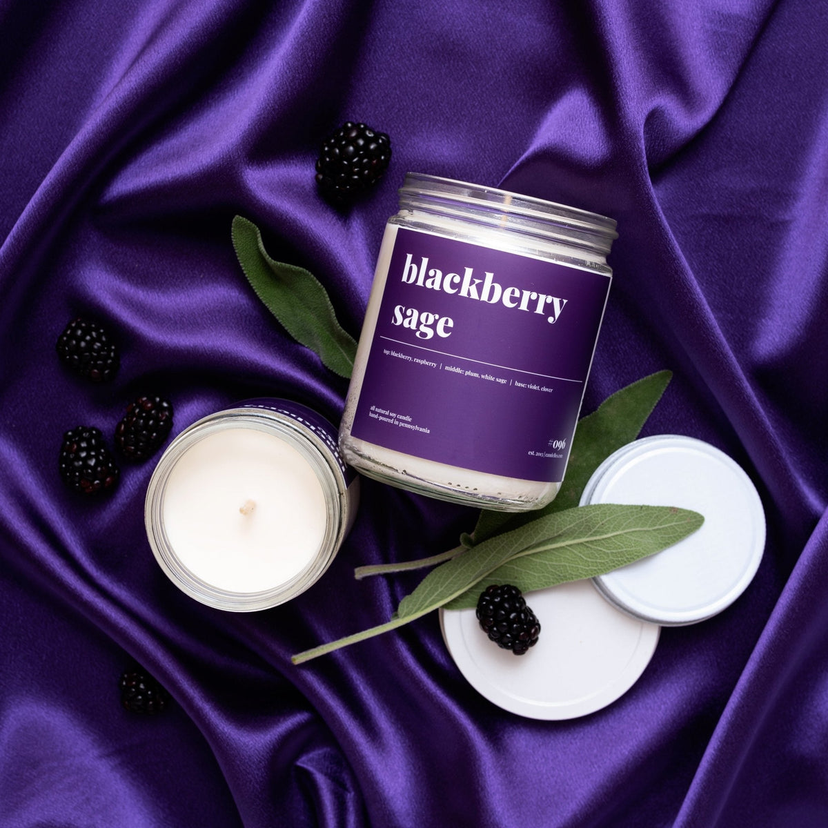 Overhead view of a blackberry sage soy candle on purple satin with an open jar, white lids, sage leaves and berries