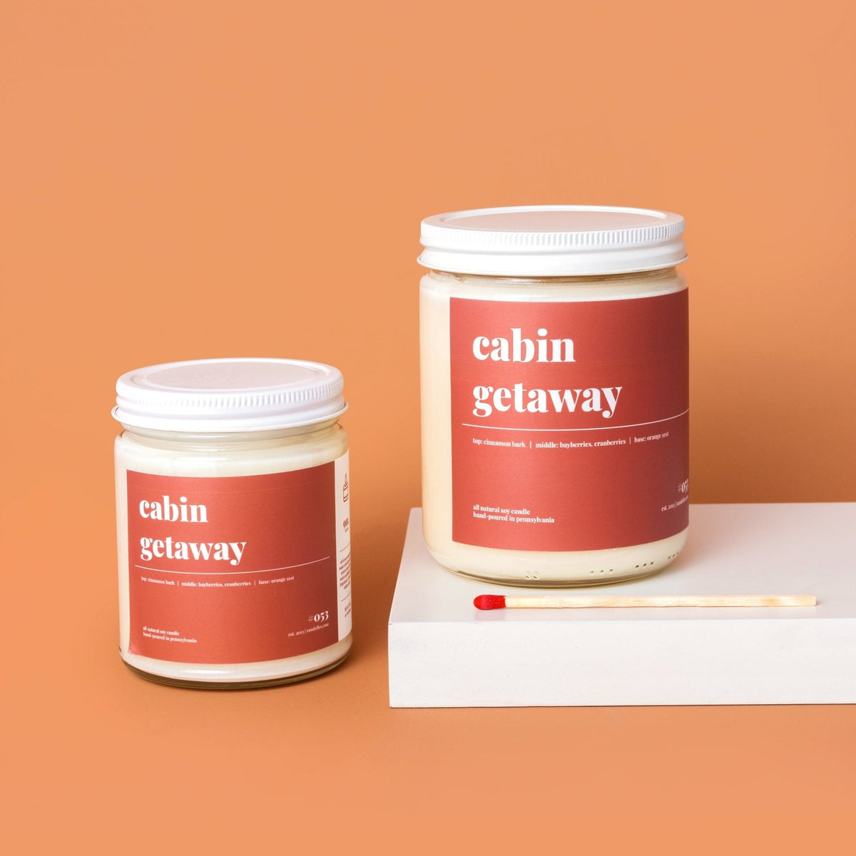 Standard and petite cabin getaway soy candles side by side on a white riser with a match on an orange background