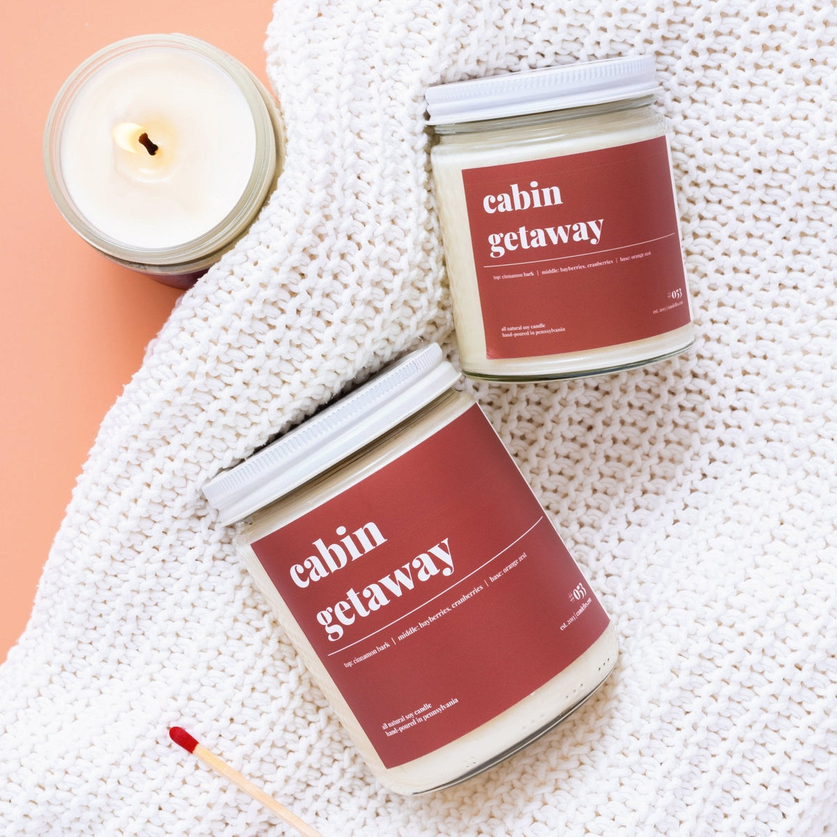 Two lidded cabin getaway candles resting on a chunky white knit blanket beside a lit jar and a red match