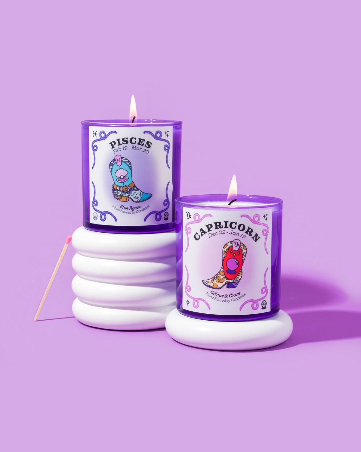 Pisces and Capricorn zodiac candles burning in purple glass tumblers on white ribbed risers against a lilac backdrop
