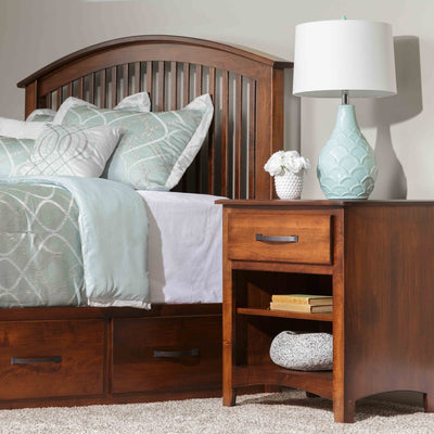 One-drawer open nightstand in brown cherry with an open shelf, black bar pull and arched base beside a spindle bed