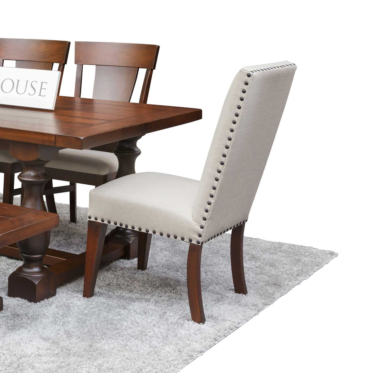 Linen parson style dining chair with nailhead trim beside a cherry trestle table and bench on a gray rug