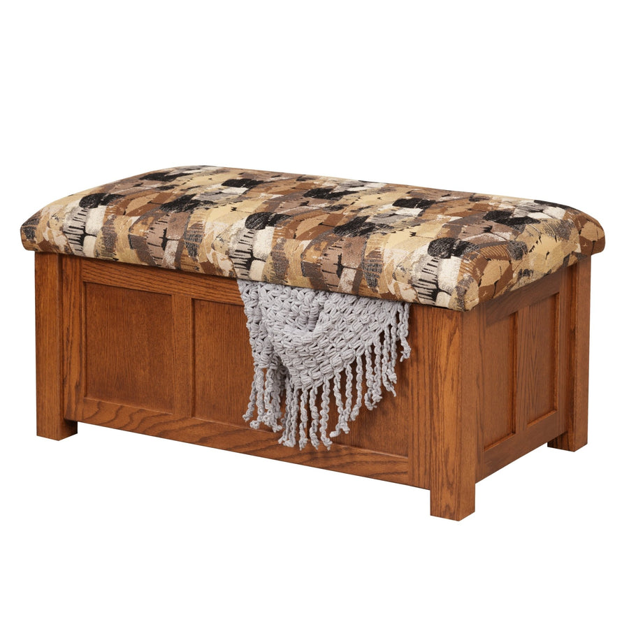 Oak storage ottoman with a padded earth tone patterned top, raised panel front and gray throw draped over