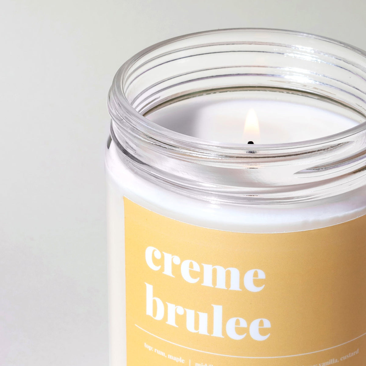 Close-up of a lit creme brulee soy candle showing the flame, melted wax pool and yellow scent label