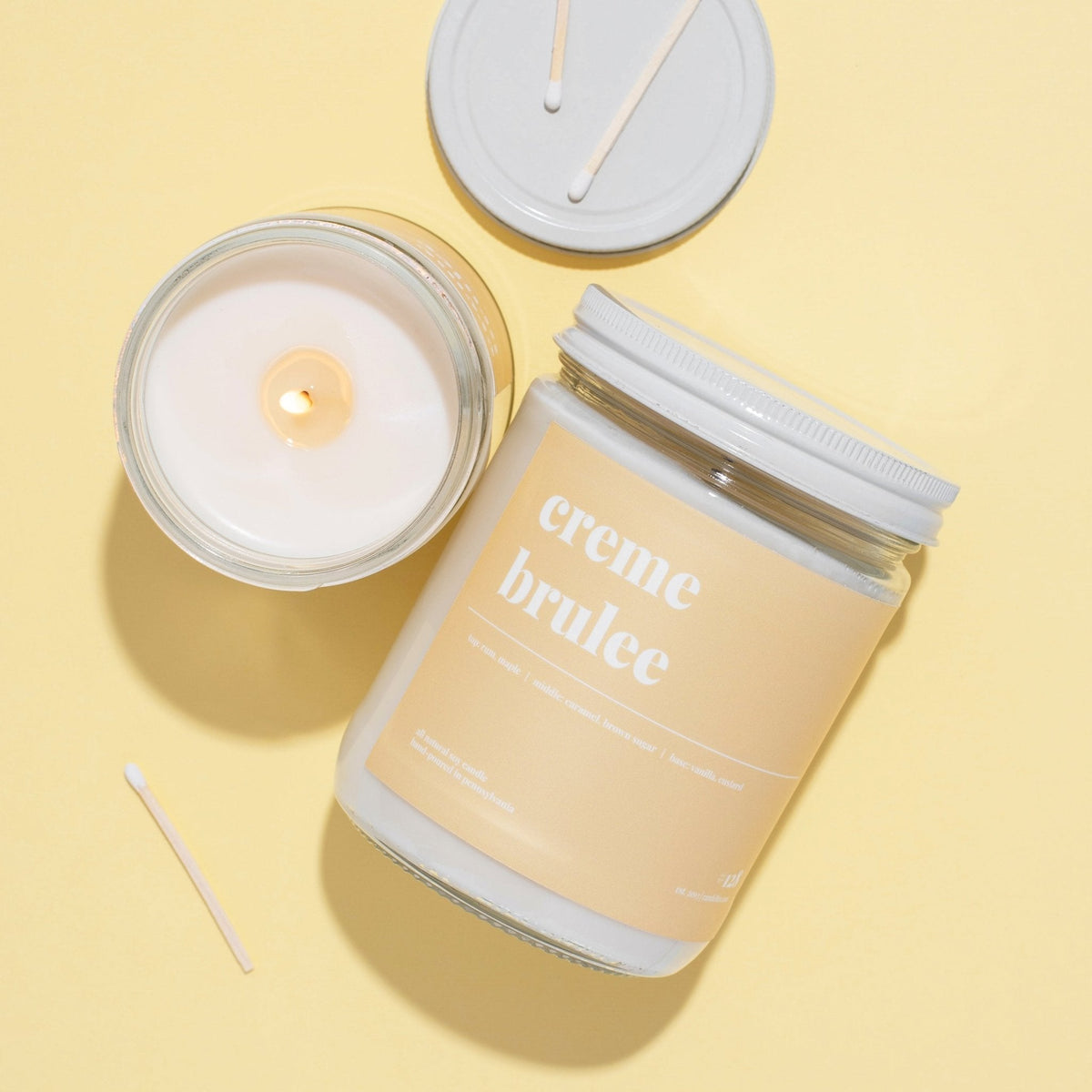 Flat lay of a creme brulee soy candle with lid, a lit open jar and matches on a butter yellow background