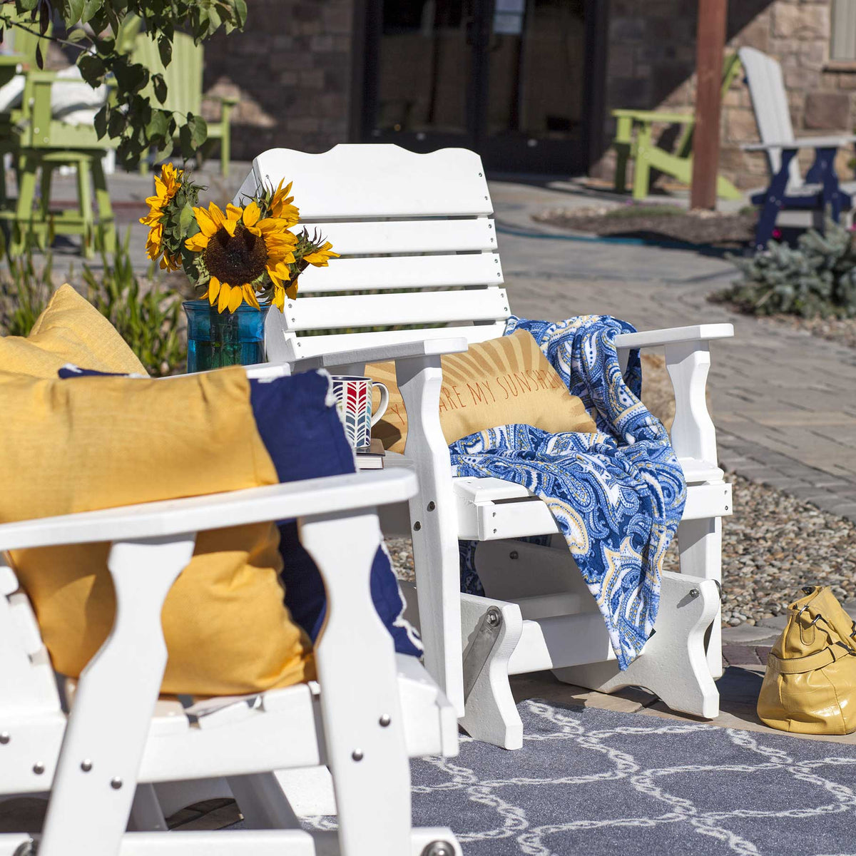 Two white poly curve back gliders with yellow and blue pillows and a paisley throw on a patio rug