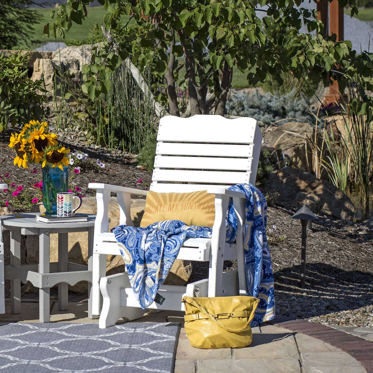 White poly curve back glider chair on a patio with a paisley throw, sunflowers and matching gray side table