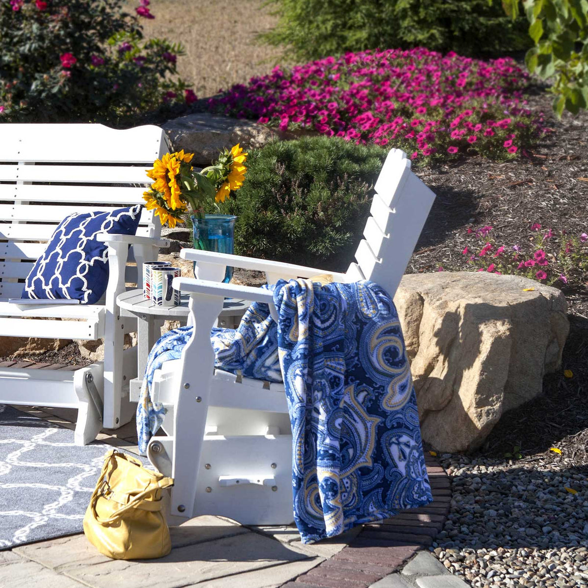 White poly glider chair and glider bench flanking a small side table with sunflowers in a landscaped garden