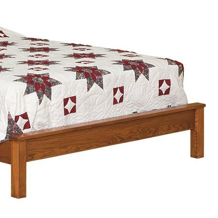 Foot of a multi panel oak bed with a low flat footboard rail and square legs under a patchwork quilt