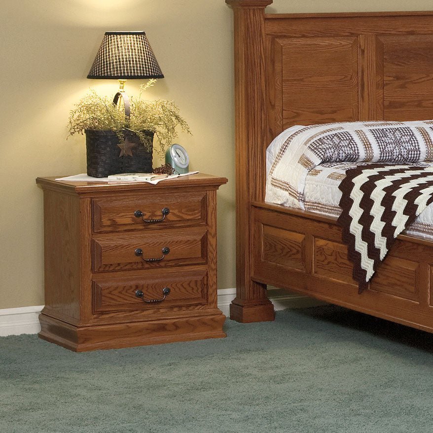 Oak three drawer nightstand styled with a lamp beside a paneled oak bed in a traditional bedroom