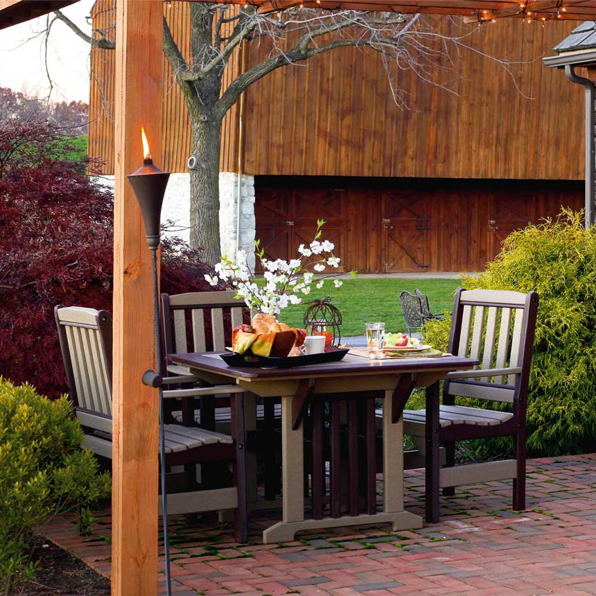 Weatherwood and cherry poly dining table for four under a pergola with tiki torch and barn in the background