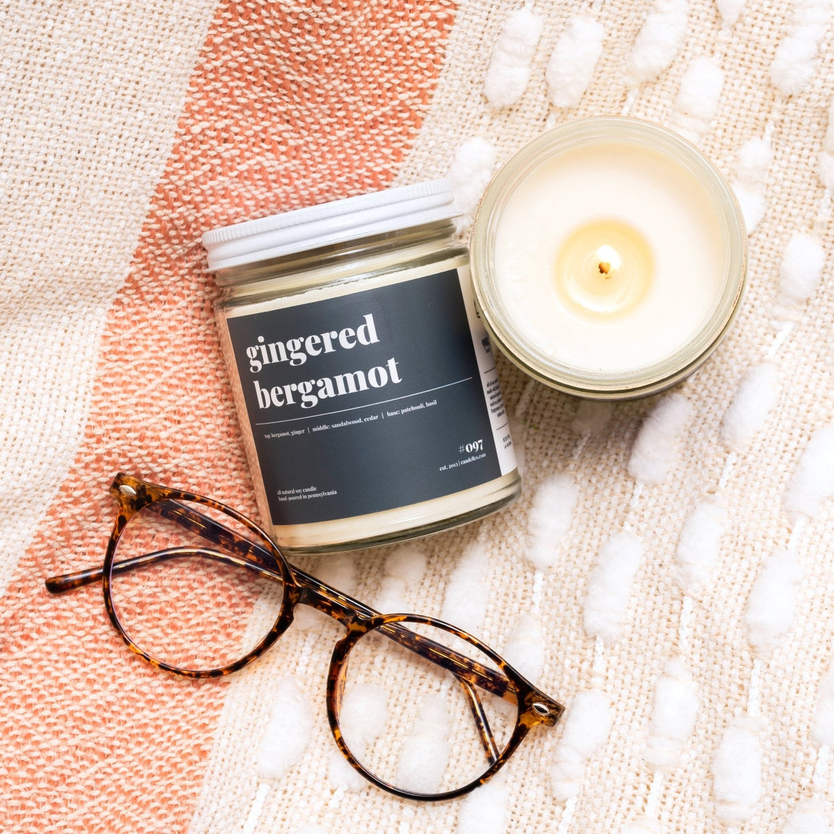 Gingered bergamot candle with black label beside a lit open jar and tortoiseshell glasses on a textured throw