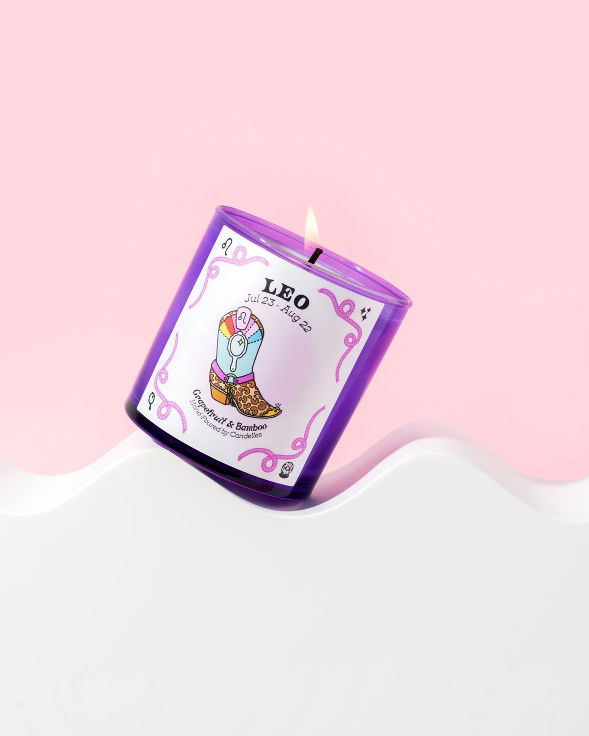 Grapefruit and bamboo Leo zodiac candle burning in a purple glass tumbler on a white wave riser against pink