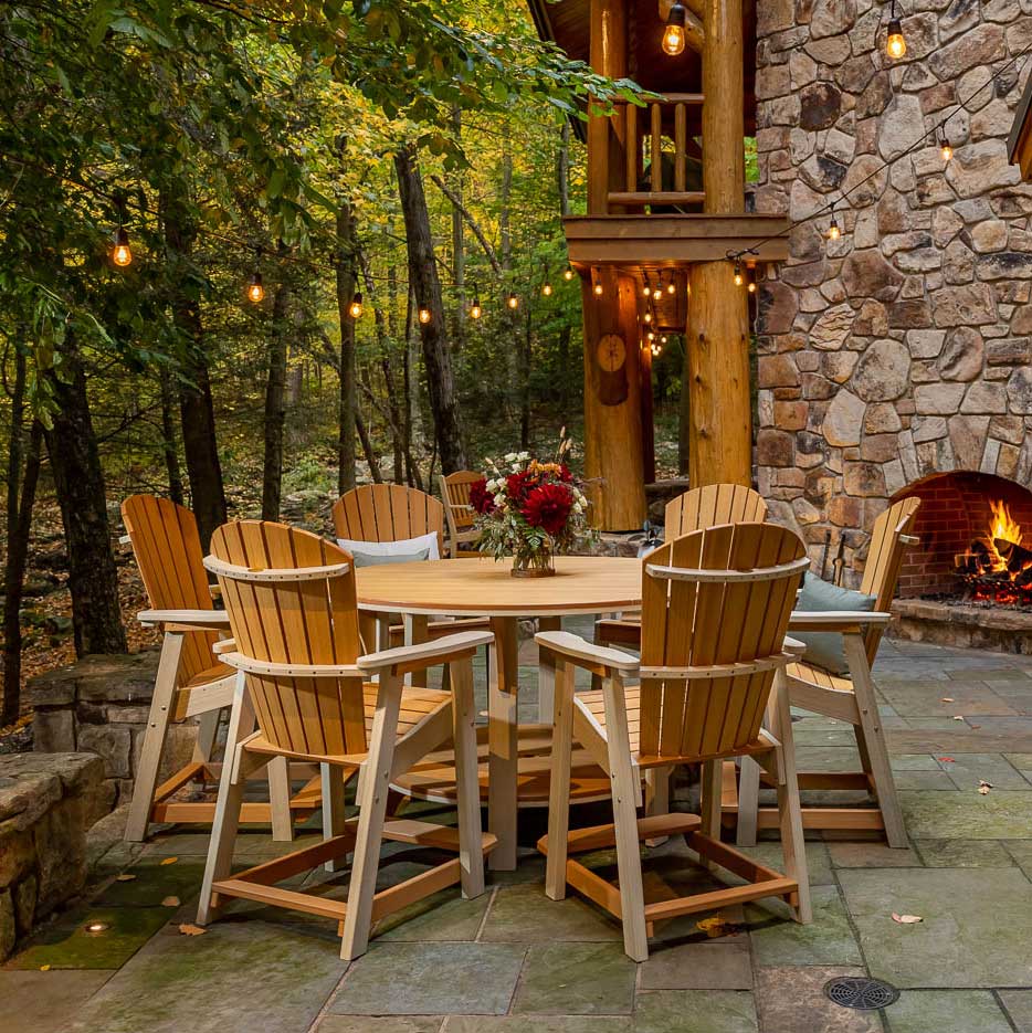 Cedar and white Adirondack counter chairs around a round poly table by a lit stone fireplace in the woods