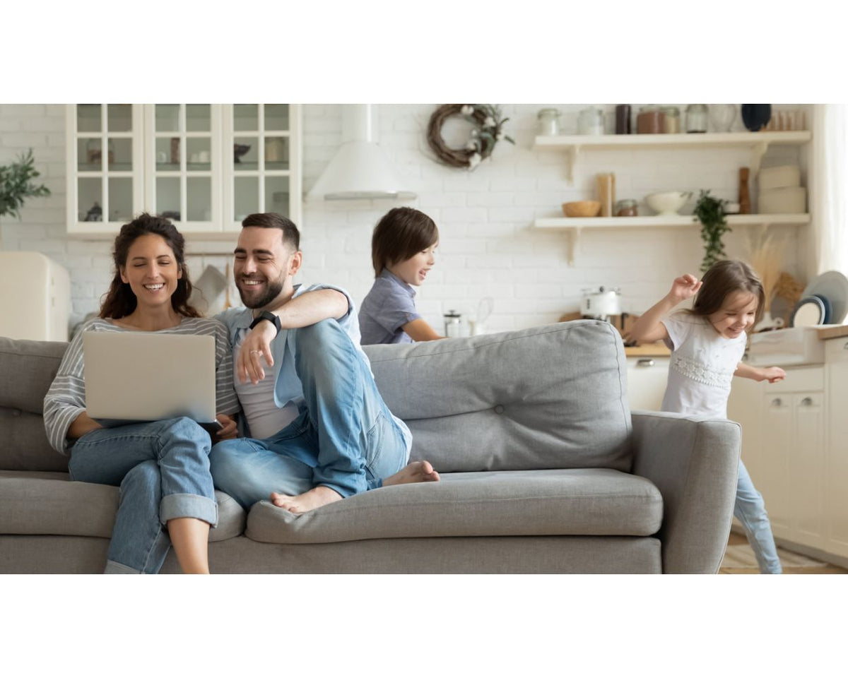Family relaxing on a gray sofa in a bright white kitchen and living space, two children playing nearby