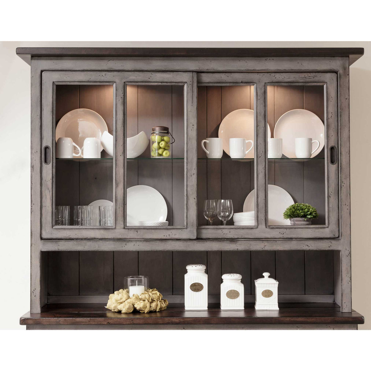 Upper section of the gray hutch showing sliding glass doors, white plates and mugs on lit shelves and canisters below