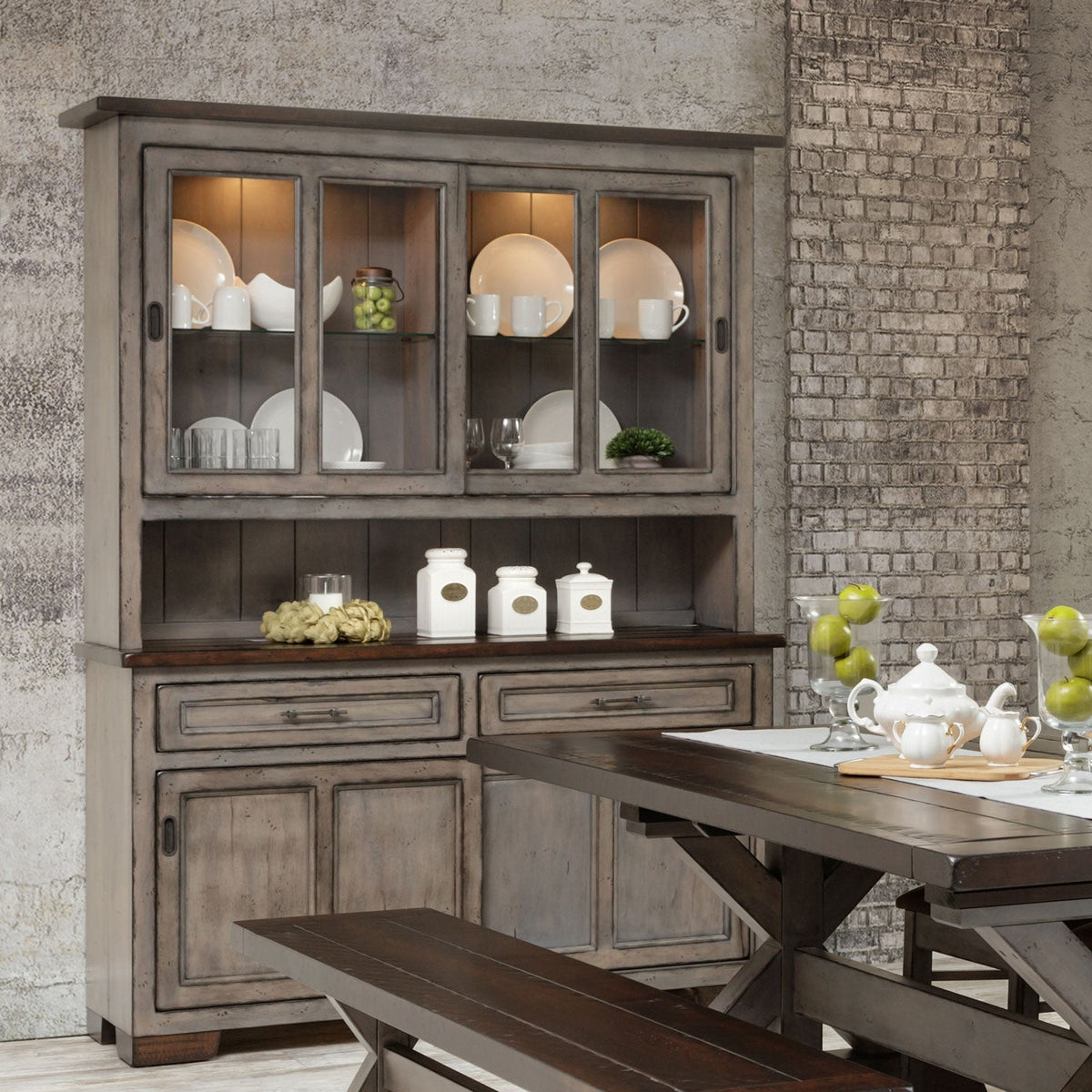 Weathered gray 63 inch hutch with lit sliding glass doors of white dishes, dark plank top and paneled base in a brick dining room