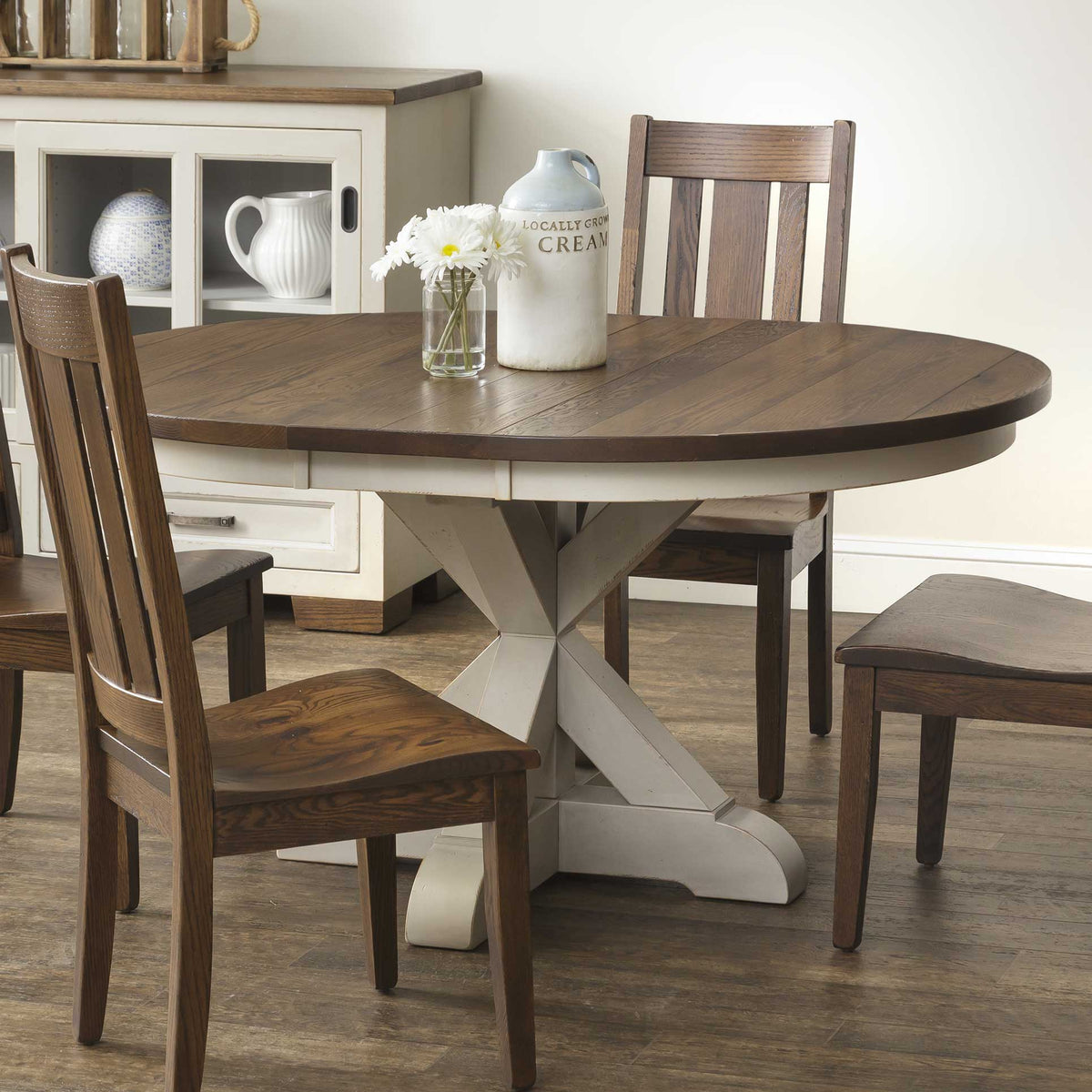 Close view of the round pedestal table edge, cream painted apron and X base with brown slat back chairs around it