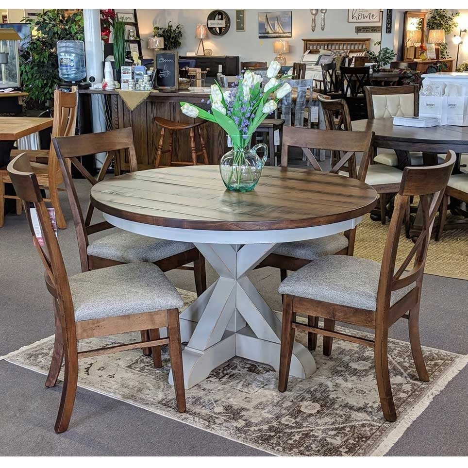 Showroom view of the round two-tone pedestal table with five X back chairs and upholstered seats on a patterned rug