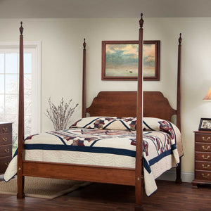 Cherry pencil post bed with four tall tapered posts, urn finials and a navy patchwork quilt in a sunny bedroom